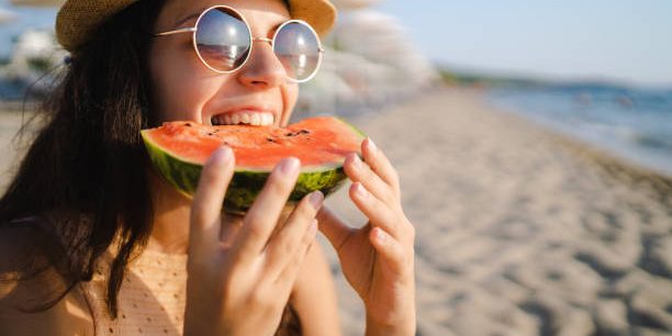 Young, happy, woman eating watermelon on the beach, wearing sunglasses.